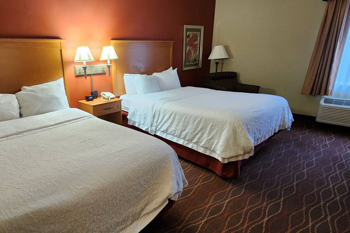 A hotel room with two beds, a nightstand with a lamp, and a wall-mounted phone.