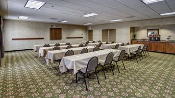 A conference room with multiple tables and chairs, carpeted floor, and a kitchenette area.