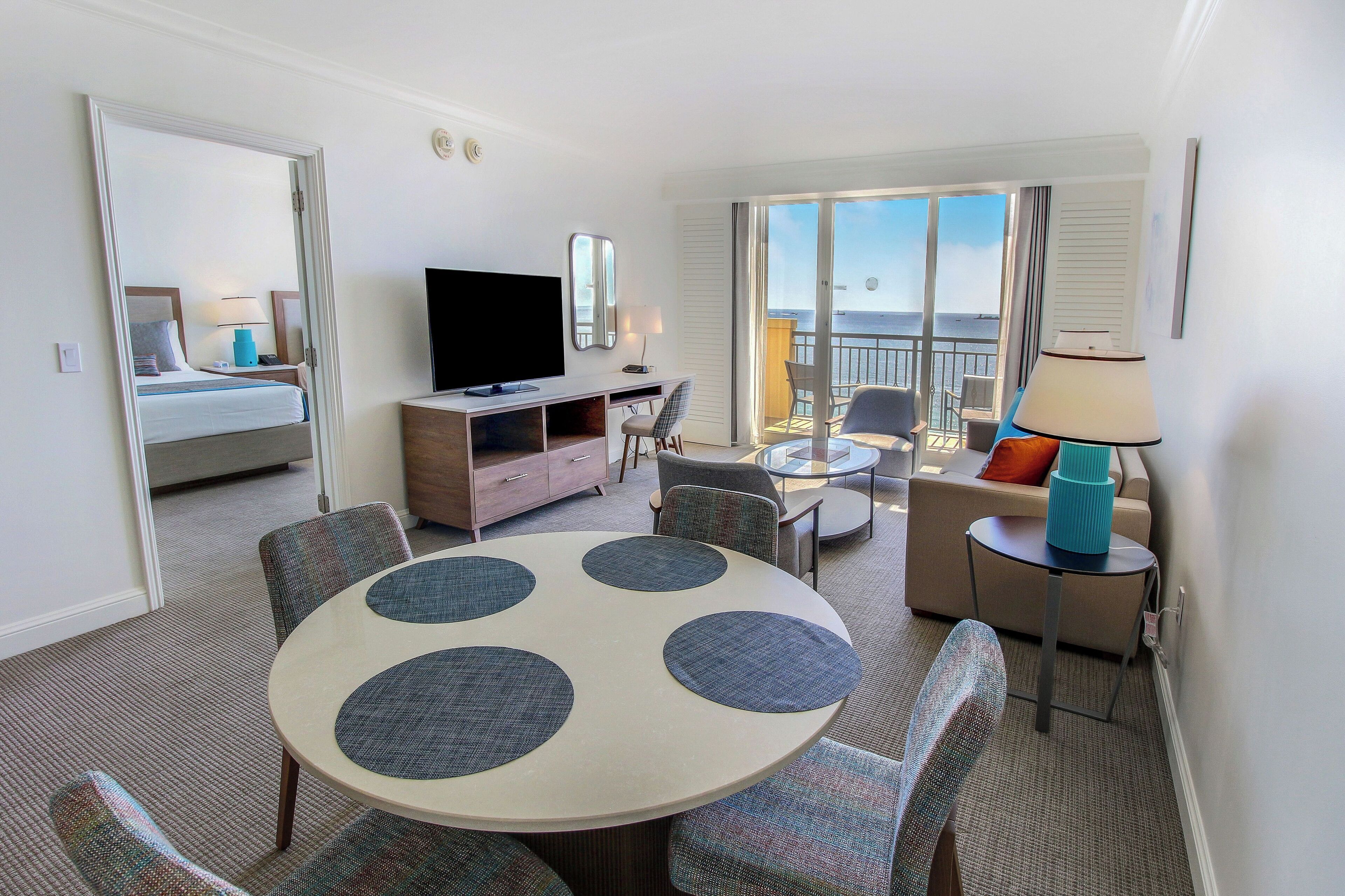 A hotel room with a balcony, a round dining table with chairs, a TV, a dresser, and a sofa.