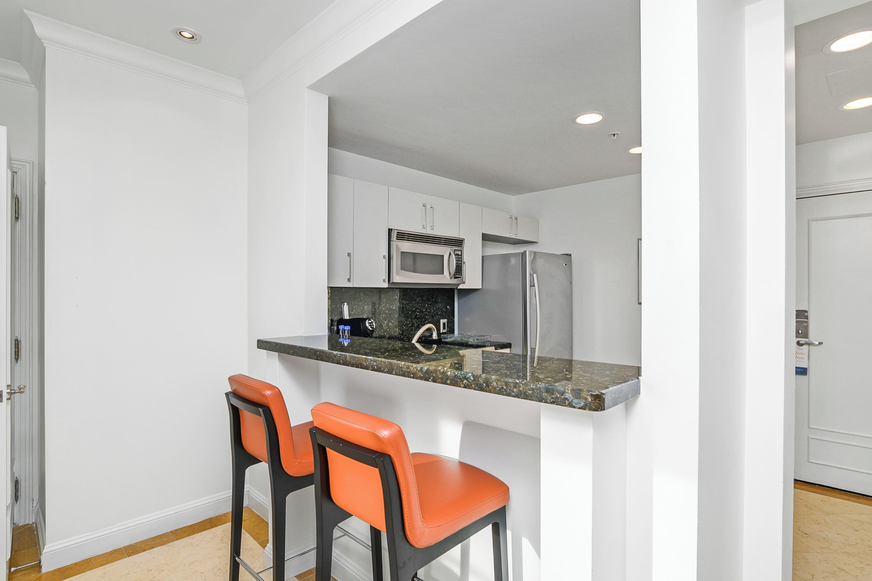 A compact kitchen with white cabinets, a stainless steel refrigerator, and a microwave. There are two orange bar stools.