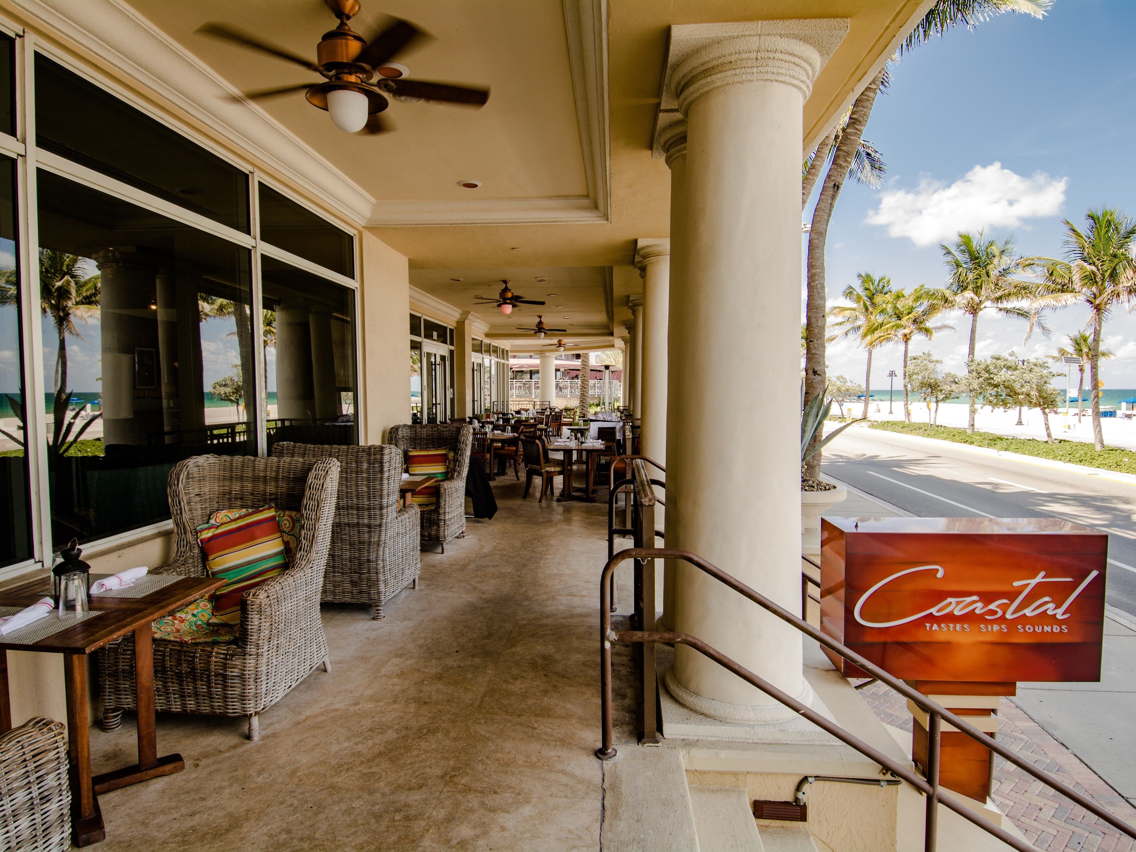 Un hall de hotel costero con muebles de mimbre, un cartel que dice "Coastal Tastes Sips Sounds" y vistas a la playa.