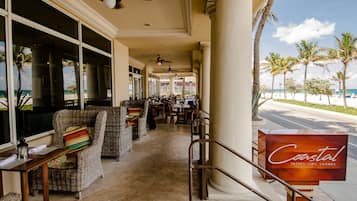 Un hall de hotel costero con muebles de mimbre, un cartel que dice "Coastal Tastes Sips Sounds" y vistas a la playa.