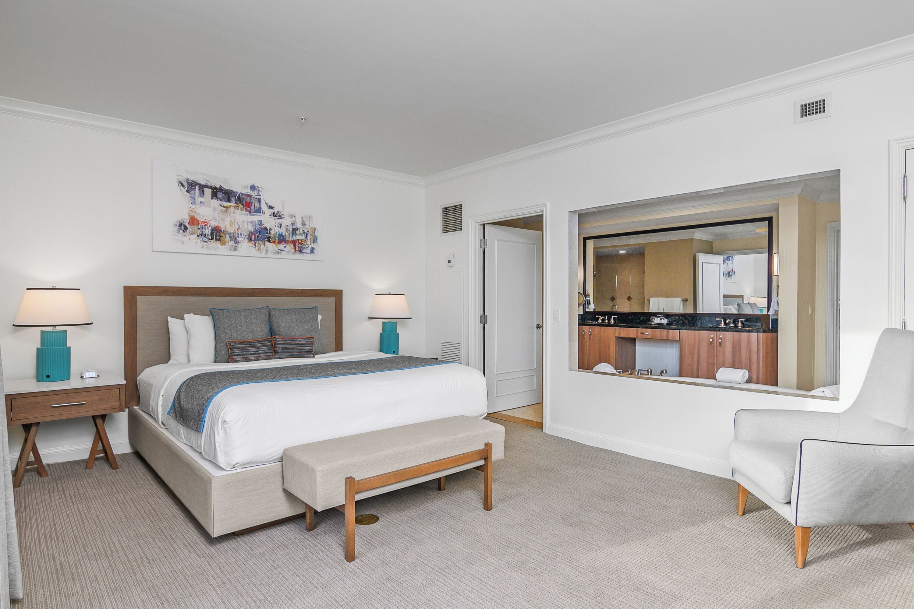 A hotel room with a large bed, bedside tables, a bench, a chair, and a view of the kitchen and bathroom through an open door.