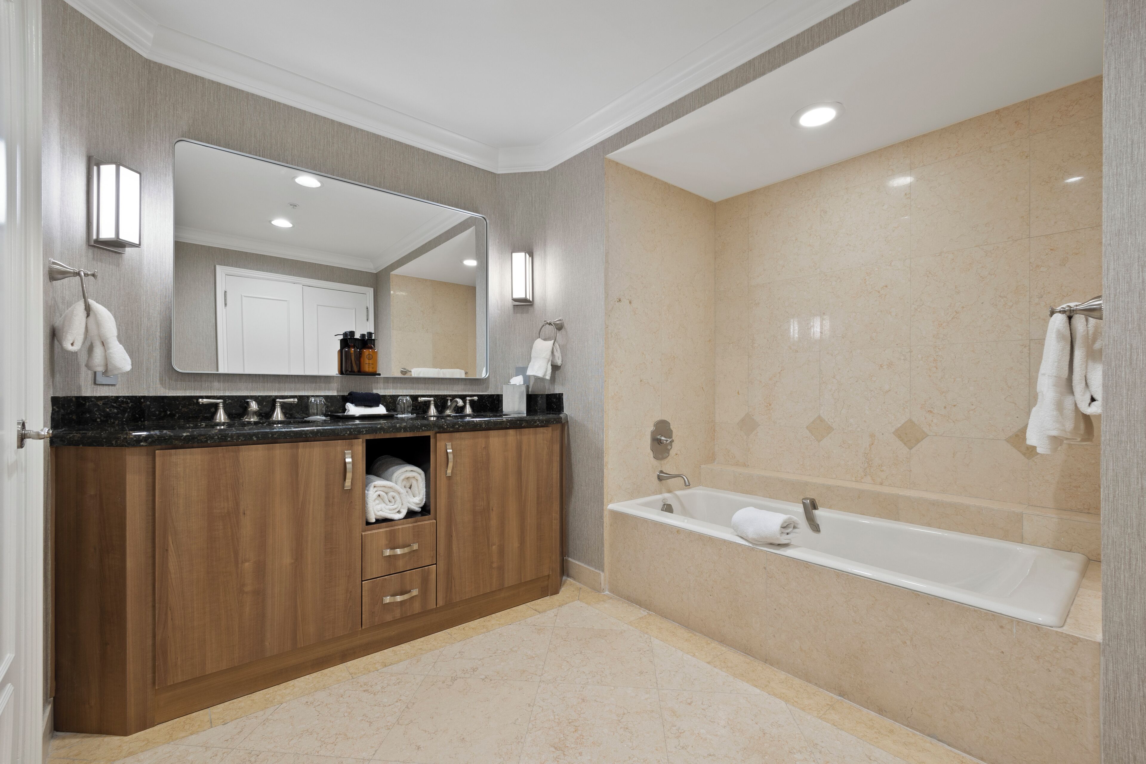 A modern bathroom with a wooden vanity, a large mirror, a bathtub, and beige tiled walls.