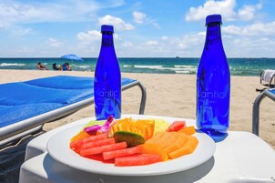 A beachside table with a plate of fruit, two blue bottles, and a beach umbrella.