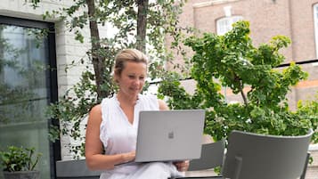 A woman sitting on a bench with a laptop, surrounded by potted plants and a modern building in the background.
