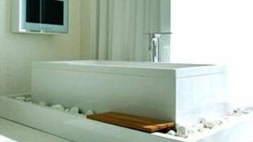 A modern bathroom with a large rectangular bathtub, a wooden bench, and a small television mounted on the wall.