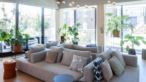 Una sala de estar moderna con un sofá gris, dos mesas negras, lámparas colgantes esféricas y ventanas amplias con plantas en macetas.