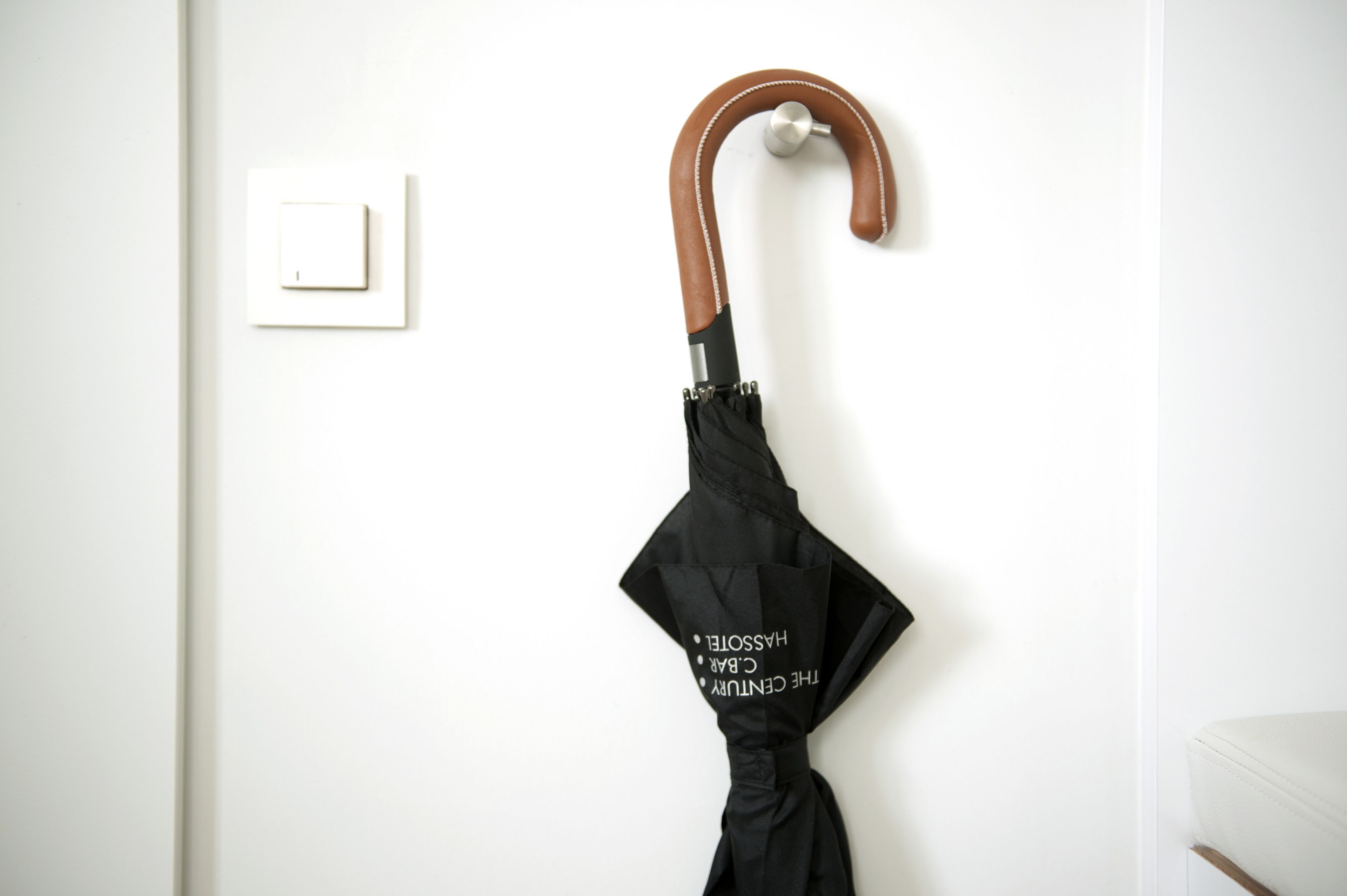 A black umbrella with a wooden handle hanging on a white wall next to a light switch.