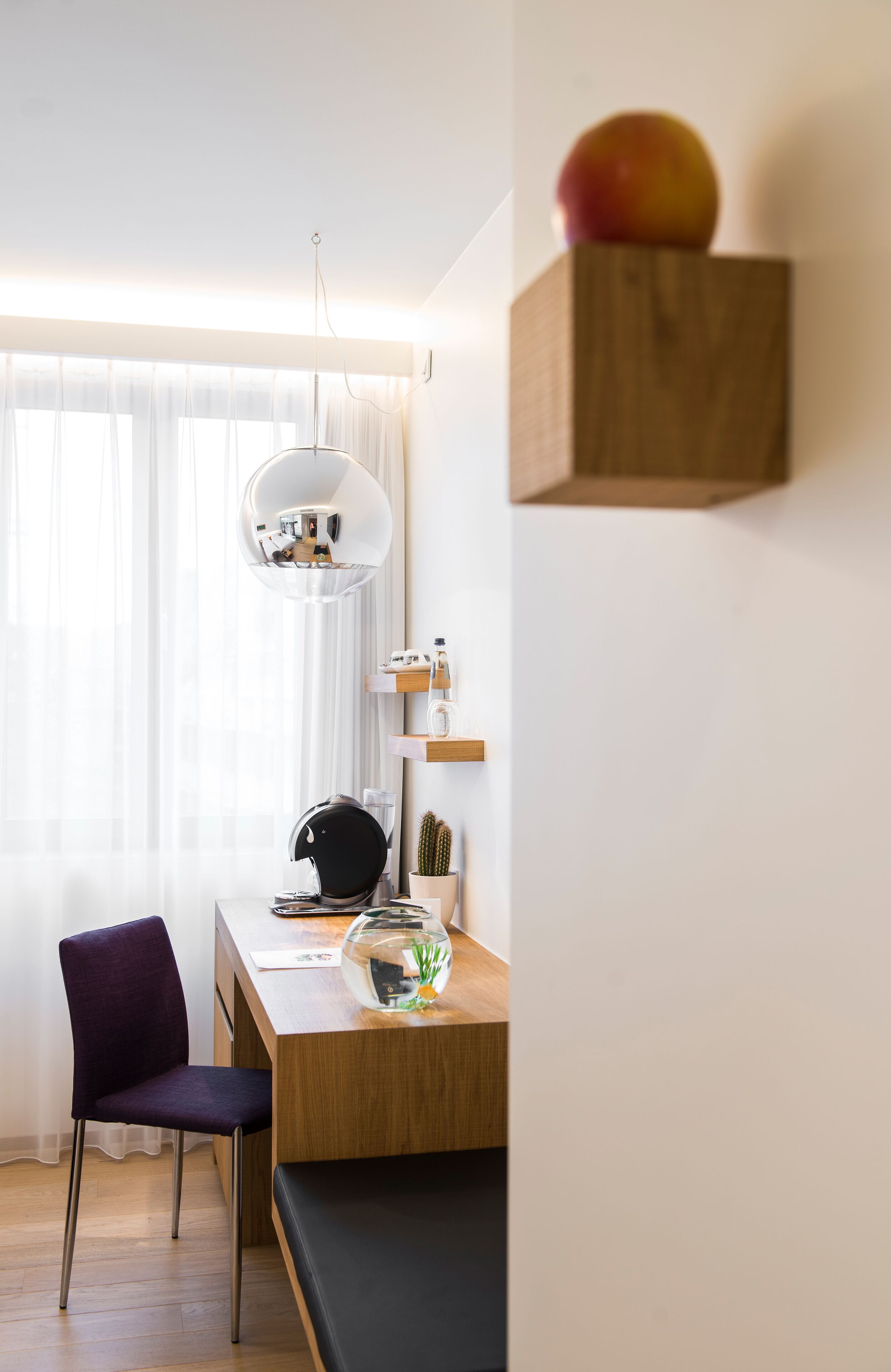 A modern, well-lit room with a wooden desk, a purple chair, a black sofa, a hanging light fixture, and a wooden shelf with a fruit.