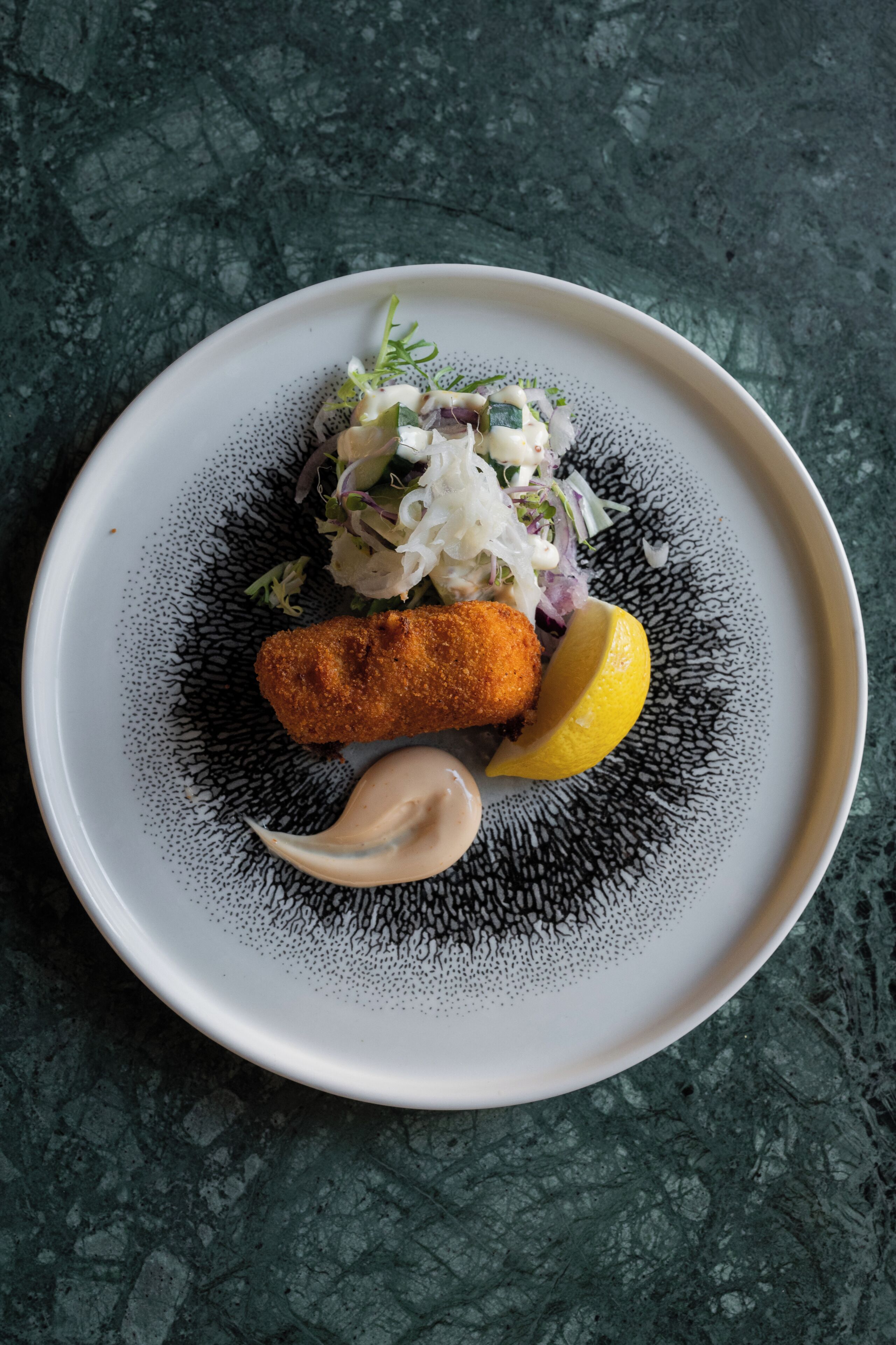 Un plato con una croqueta, ensalada y una cuña de limón sobre un plato con textura.