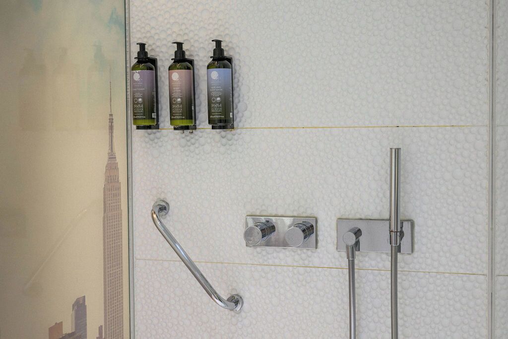 A bathroom with a handrail, a showerhead, and three bottles of shampoo on a textured wall.