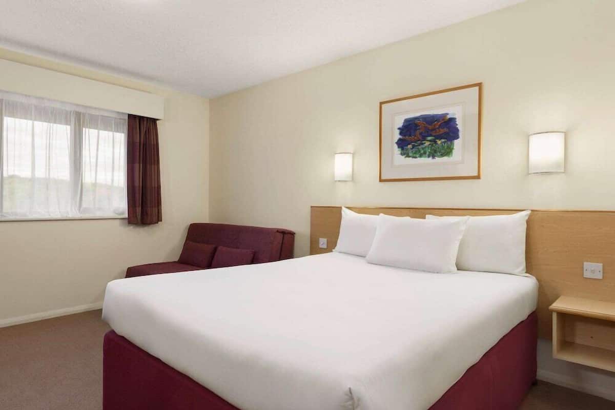 A hotel room with a large bed, a desk, a chair, a window with curtains, and a framed picture on the wall.