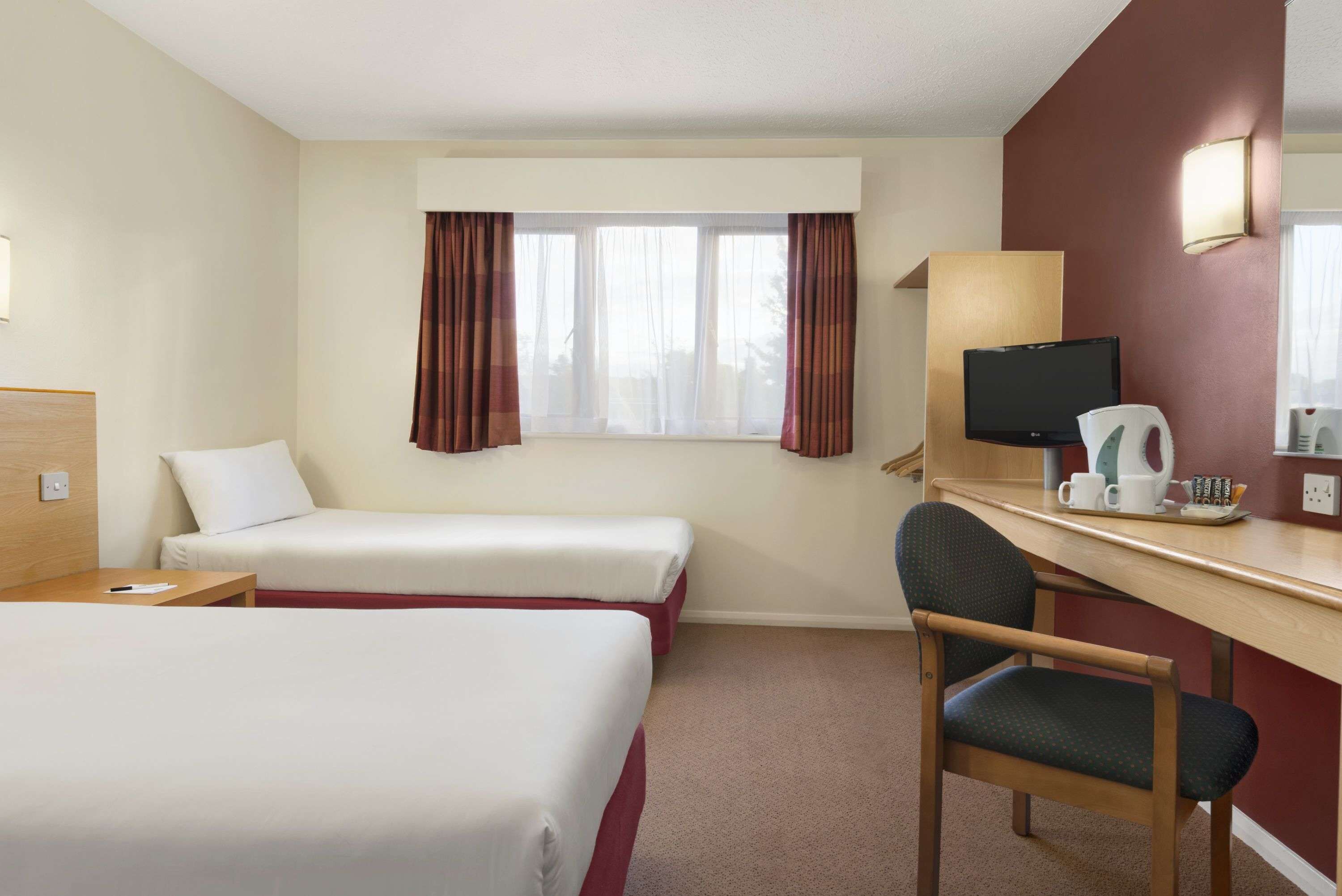 A hotel room with two single beds, a desk with a chair, a television, and a window with curtains.