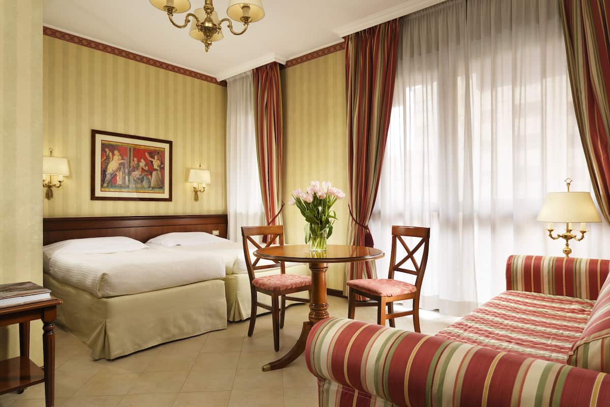 Una camera d'albergo con un letto, un divano, un tavolo da pranzo con sedie, un lampadario e un quadro appeso al muro.