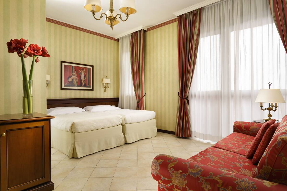 Camera d'albergo con due letti, un divano rosso, un lampadario e carta da parati a righe.