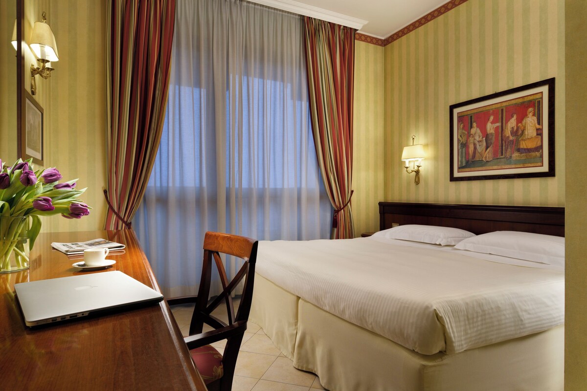 Una habitación de hotel con una cama, un escritorio con una computadora portátil, una silla, un jarrón con flores, un cuadro en la pared y cortinas.