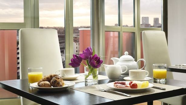 Une table de petit-déjeuner avec des croissants, des fruits et du jus, placée devant une grande fenêtre offrant une vue sur la ville.