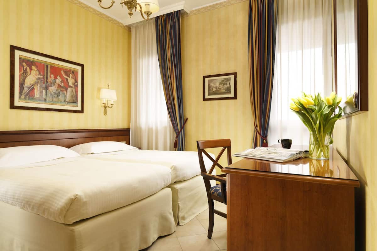 Una camera d'albergo con due letti, una scrivania con una sedia, una lampada e un vaso con fiori.