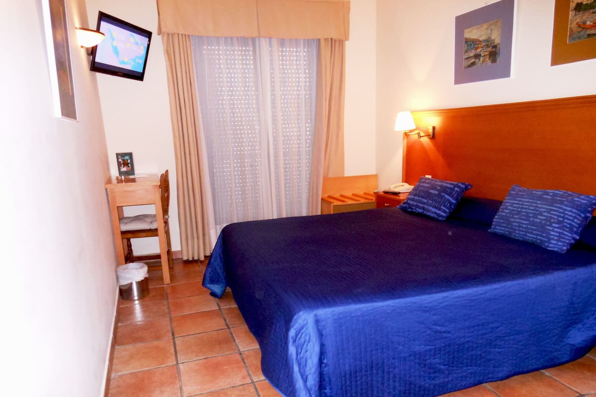 A hotel room with a large bed, a desk, a chair, a nightstand, a lamp, a TV on the wall, and a window with curtains.