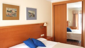 Habitación de hotel con cabecera de madera, cama con almohadas azules, mesita de noche con lámpara y dos cuadros enmarcados en la pared.