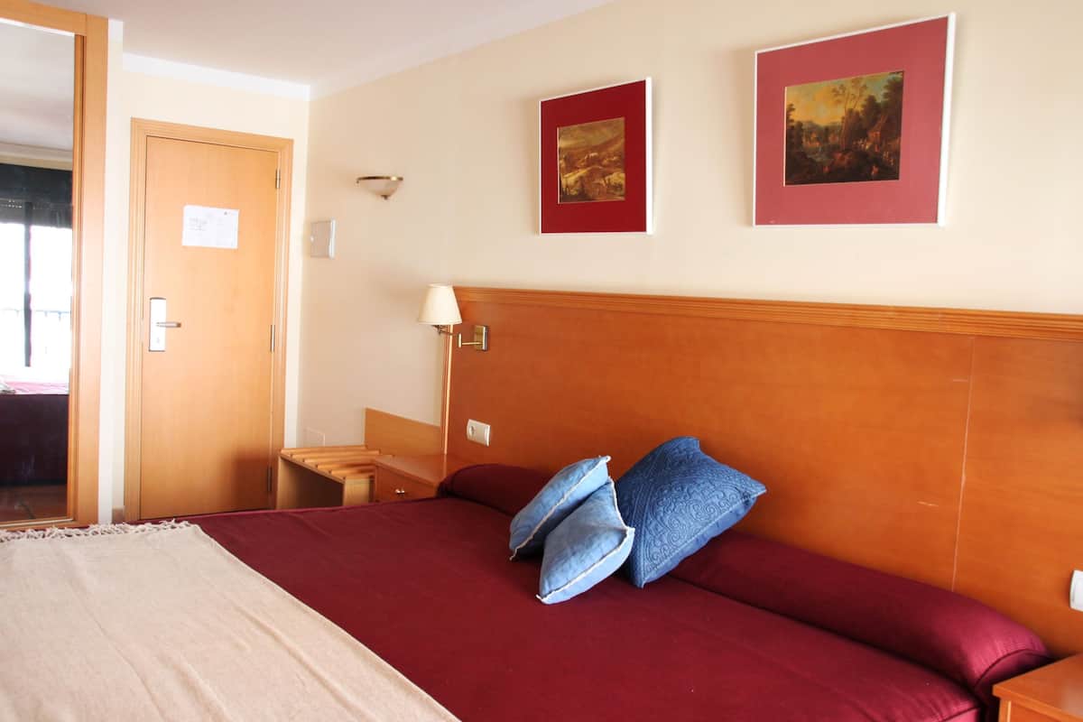 A hotel room with a wooden headboard, a bed with red bedding and blue pillows, a nightstand, a lamp, and two framed pictures on the wall.