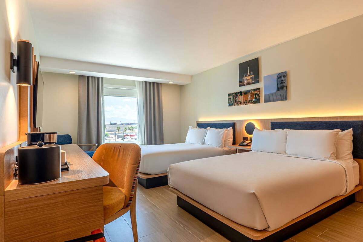 Ett hotellrum med två sängar, ett skrivbord, en stol och ett stort fönster med gardiner.