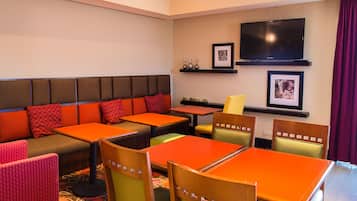 A hotel room with a seating area featuring orange tables and chairs, a sofa with red cushions, and a television mounted on the wall.
