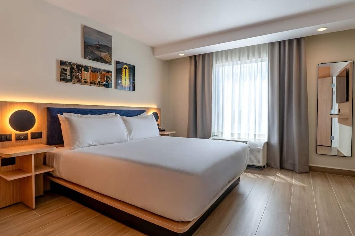 Ett modernt hotellrum med en stor säng, ett sängbord med en lampa, en stor spegel och konstverk på väggen.