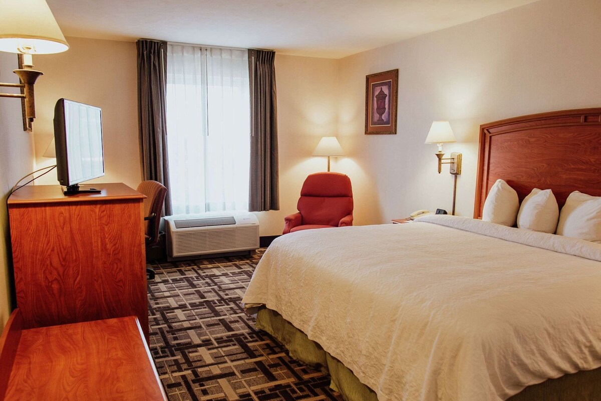 Ein Hotelzimmer mit einem Bett, einem Schreibtisch, einem Sessel, einem Fernseher, einer Lampe und einem Fenster mit Vorhängen.