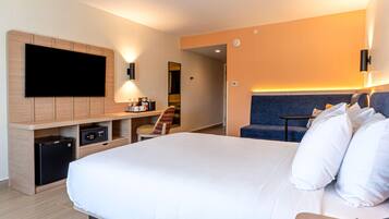 Een moderne hotelkamer met een groot bed, een flatscreen televisie bevestigd aan een houten paneel, een bureau met een computer en een lamp, en een zithoek met een bank en kussens.