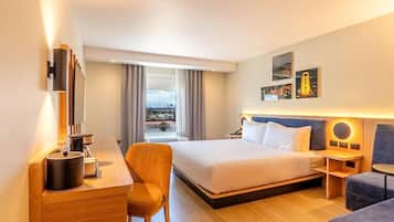 A hotel room with a bed, a desk with a lamp, a chair, a bedside table, and a view of the city through large windows.