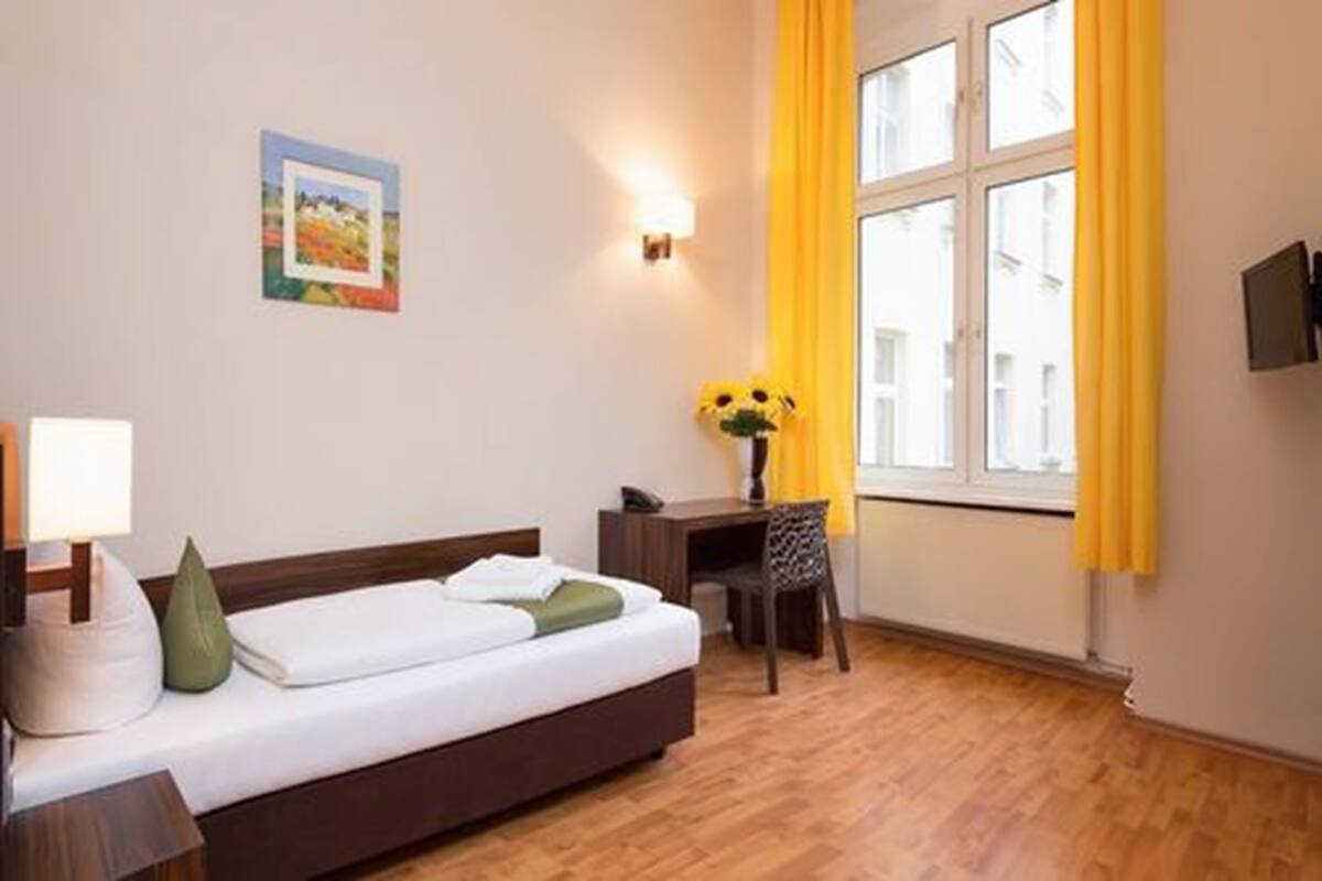 A hotel room with a bed, a desk, a chair, a lamp, a painting, and a window with yellow curtains.
