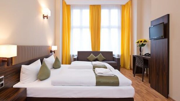 A hotel room with a large bed, a desk, a television, and a window with yellow curtains. - Hotel Abendstern (Berlin)