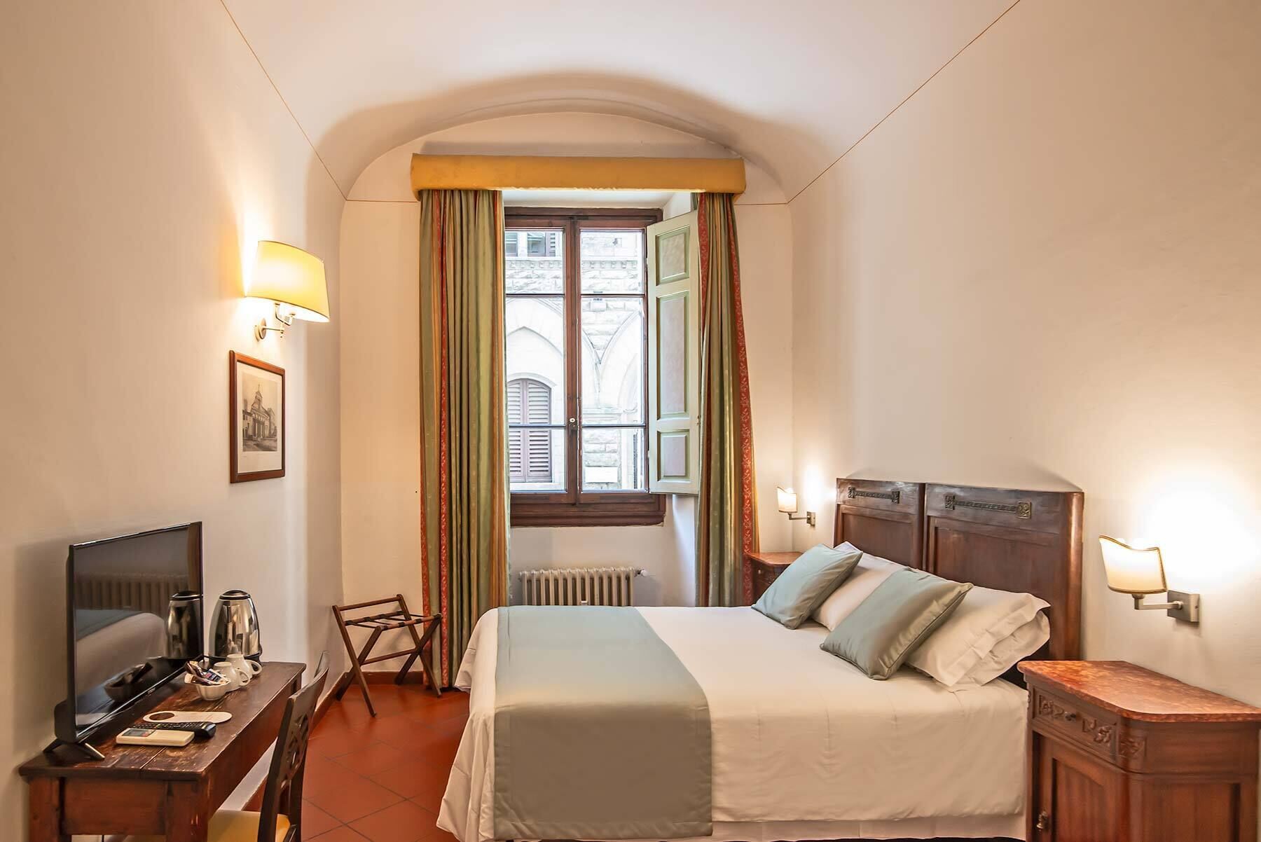 A hotel room with a bed, a desk with a TV, a chair, a radiator, and a window with curtains.
