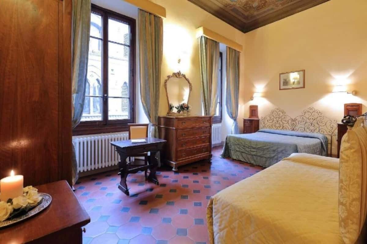 A hotel room with a wooden dresser, a desk with a candle, a bed with a green bedspread, and a window with curtains.