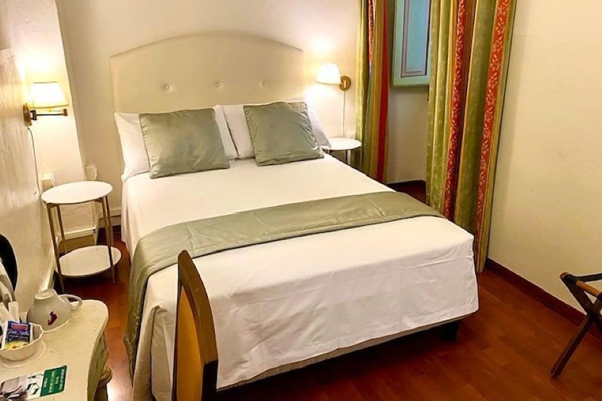 A neatly made bed with a white headboard, two bedside tables with lamps, and a small table with a kettle and a cup.