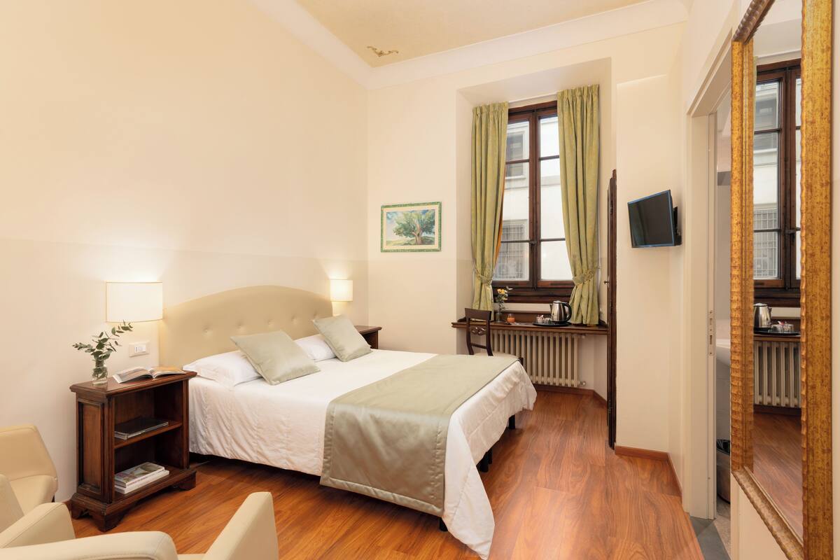 A hotel room with a bed, bedside table, lamp, chair, and a view of the outside through a window.