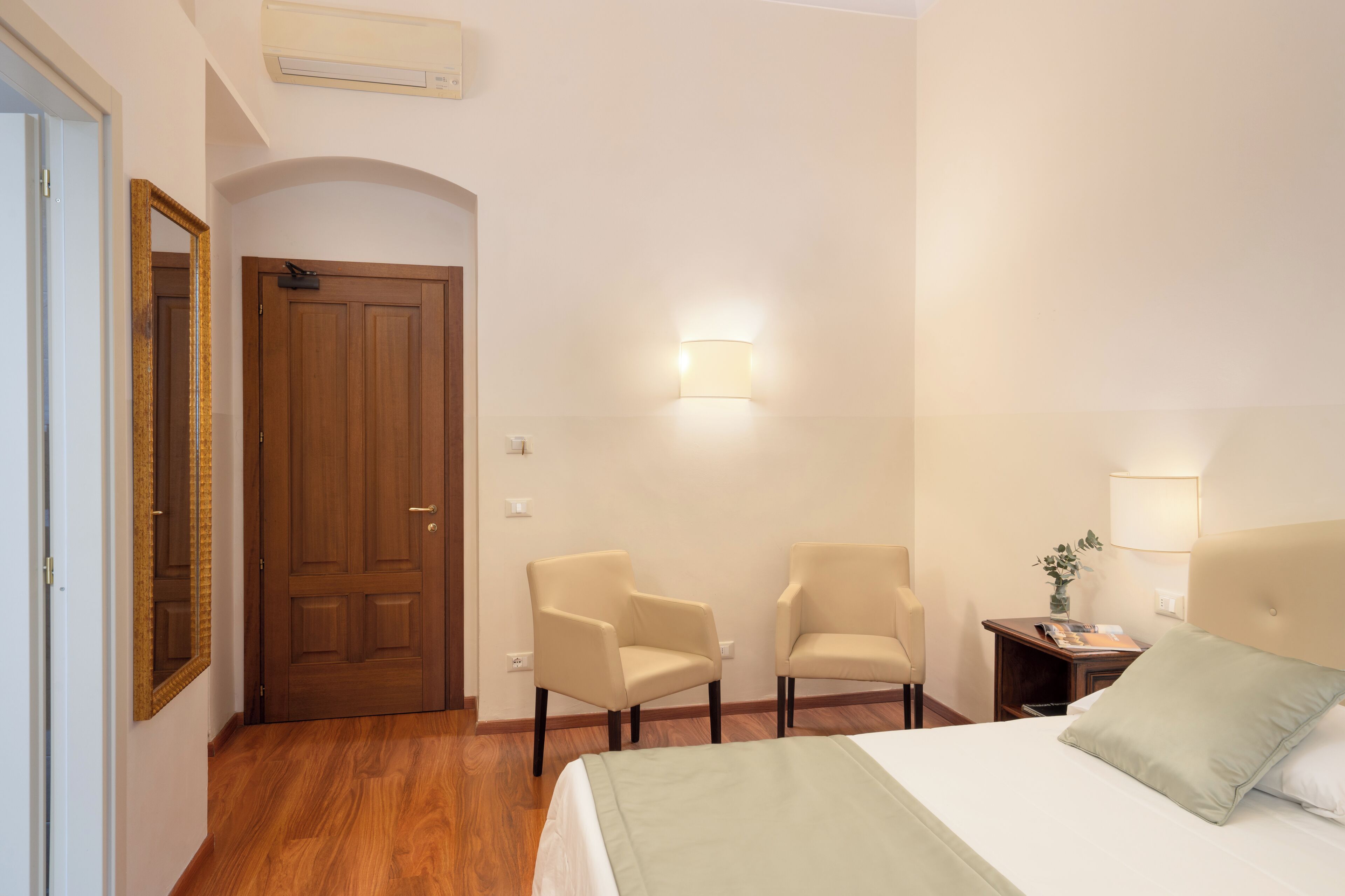 Una camera d'albergo con una porta in legno, due poltrone, un letto con cuscino verde, un comodino con una lampada e una luce a parete.