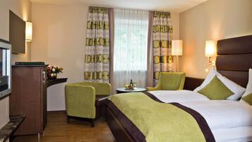 A hotel room with a bed, a desk, a chair, a small table with a bottle and fruit, and curtains with a geometric pattern.
