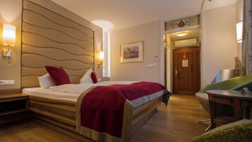 A hotel room with a large bed, a wooden headboard, a red throw blanket, a green patterned armchair, and a painting on the wall.