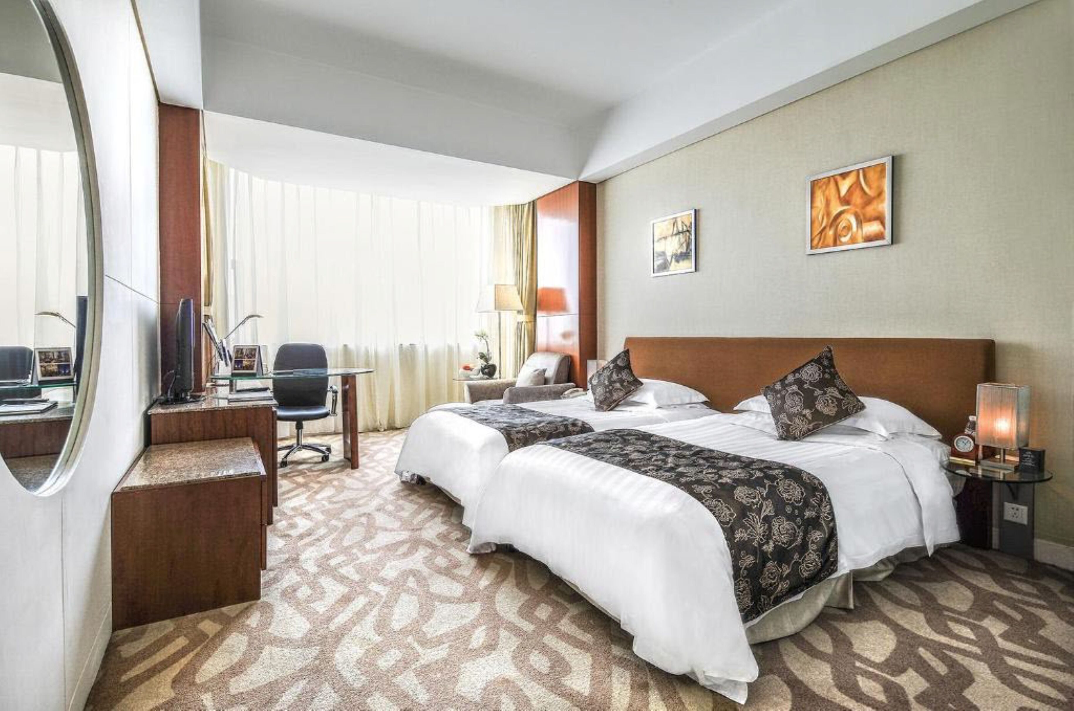 A hotel room with two beds, a desk with a computer, a chair, a lamp, and artwork on the wall.