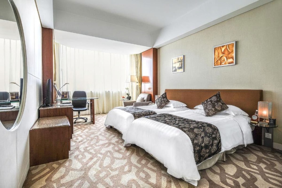 Une chambre d’hôtel avec deux lits, un bureau avec un ordinateur, une chaise, une lampe et des œuvres d’art accrochées au mur.
