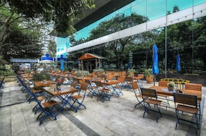 Un café en plein air, avec des tables et des chaises, entouré de verdure et bordé d’un bâtiment moderne doté de grandes fenêtres.