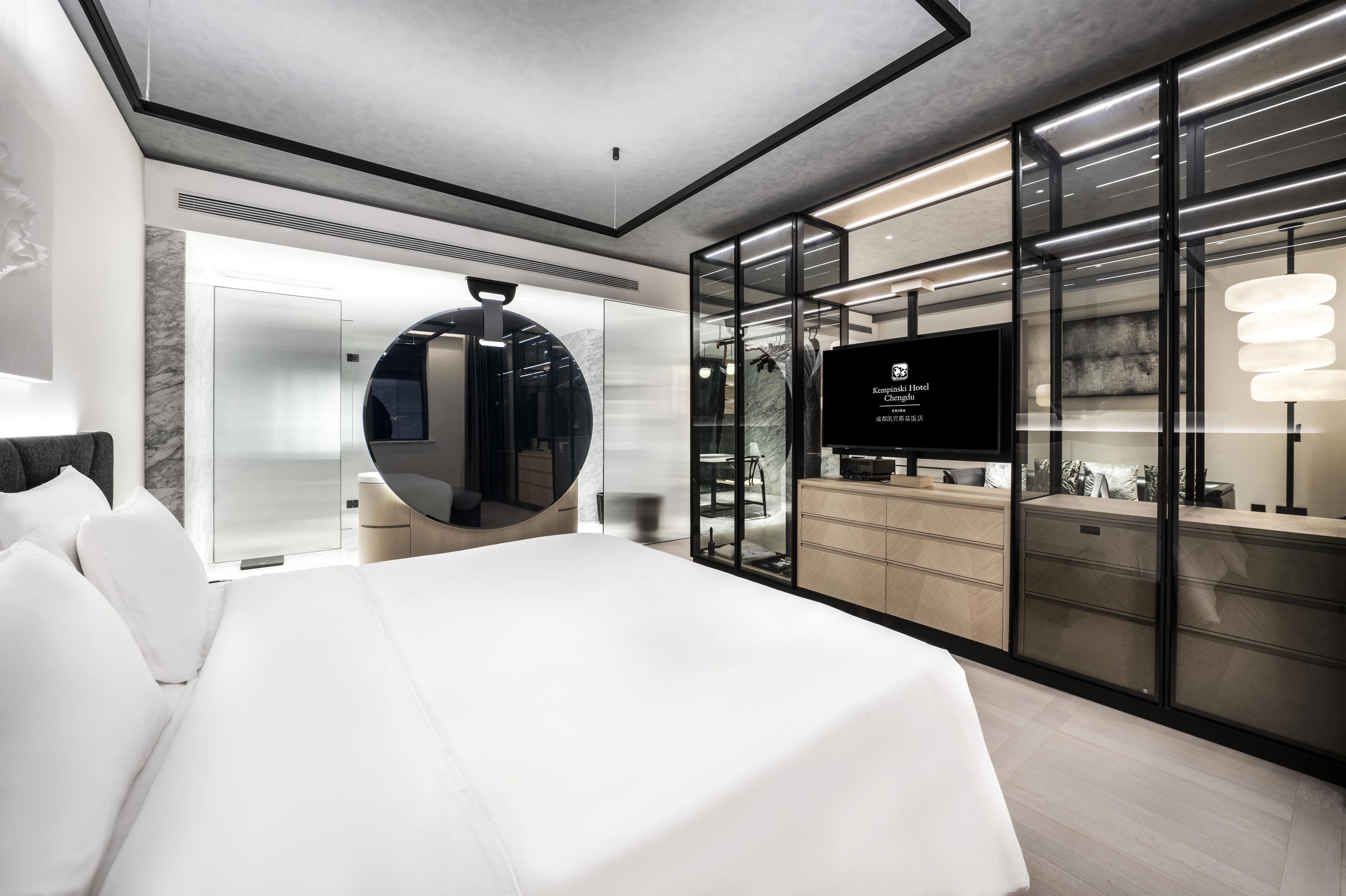 A modern hotel room with a large bed, a round mirror, a television, and a minibar.