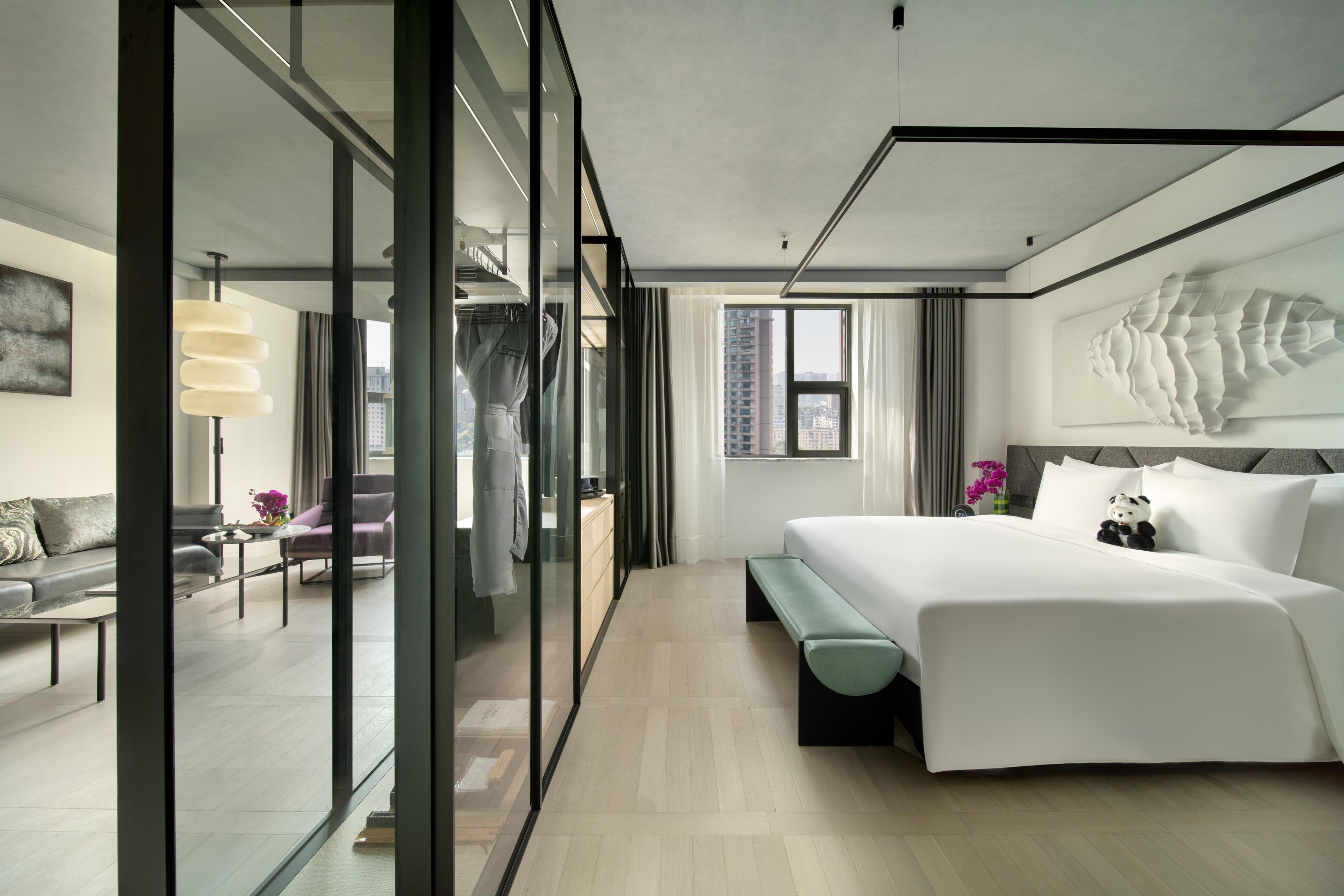 A modern hotel room with a large bed, a seating area with a sofa and armchair, a glass partition, and a view of the cityscape.