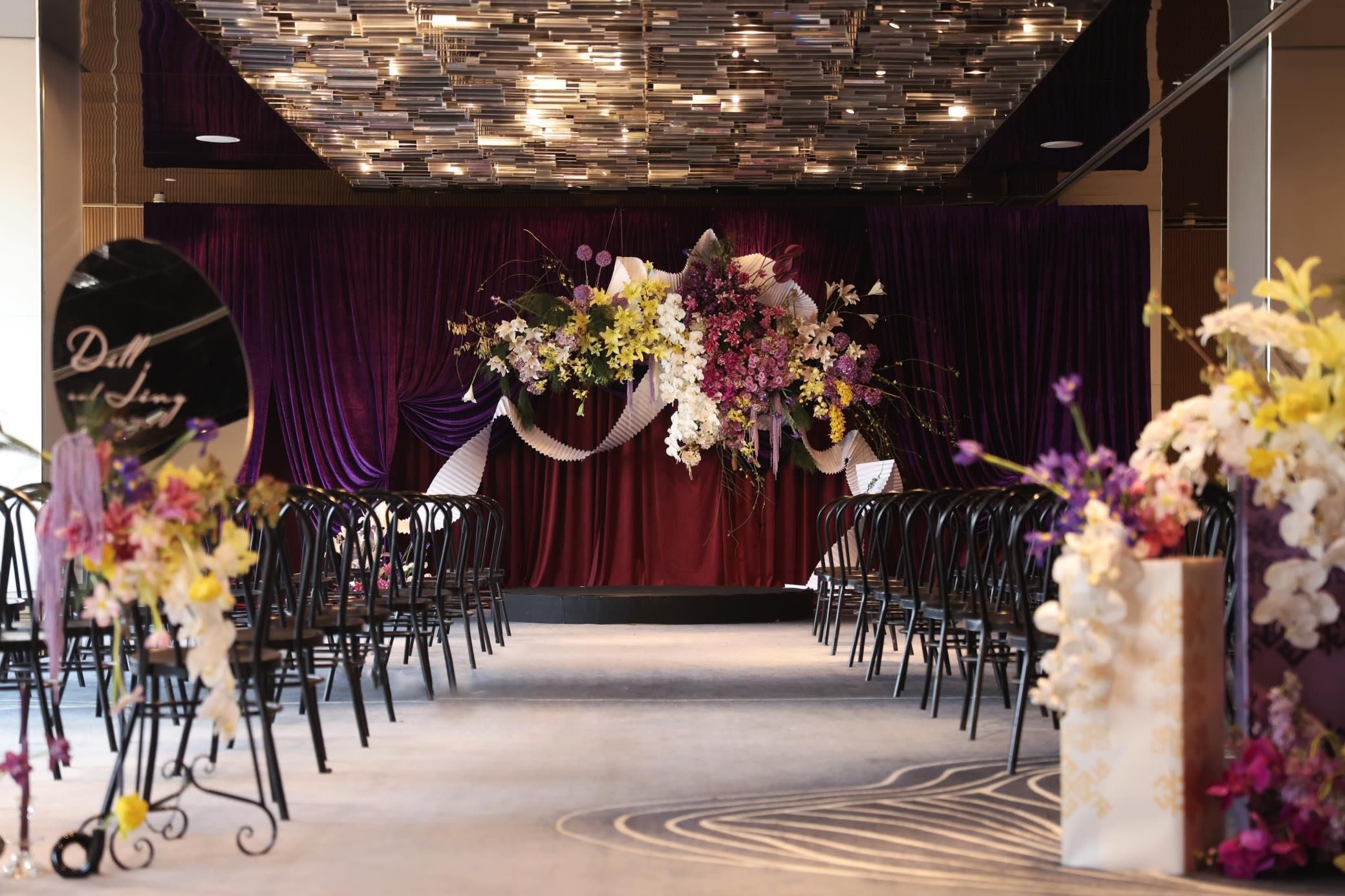 A venue with rows of black chairs arranged for an event, a stage with a red curtain backdrop, and floral arrangements.