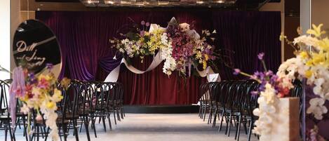 A venue with rows of black chairs arranged for an event, a stage with a red curtain backdrop, and floral arrangements.