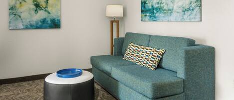 A room with a teal sofa, a patterned pillow, a blue bowl on a round ottoman, and two abstract paintings on the walls.