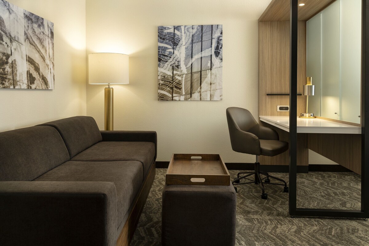 A modern hotel room with a gray sofa, a desk with a chair, a wooden side table, and two wall-mounted artworks.